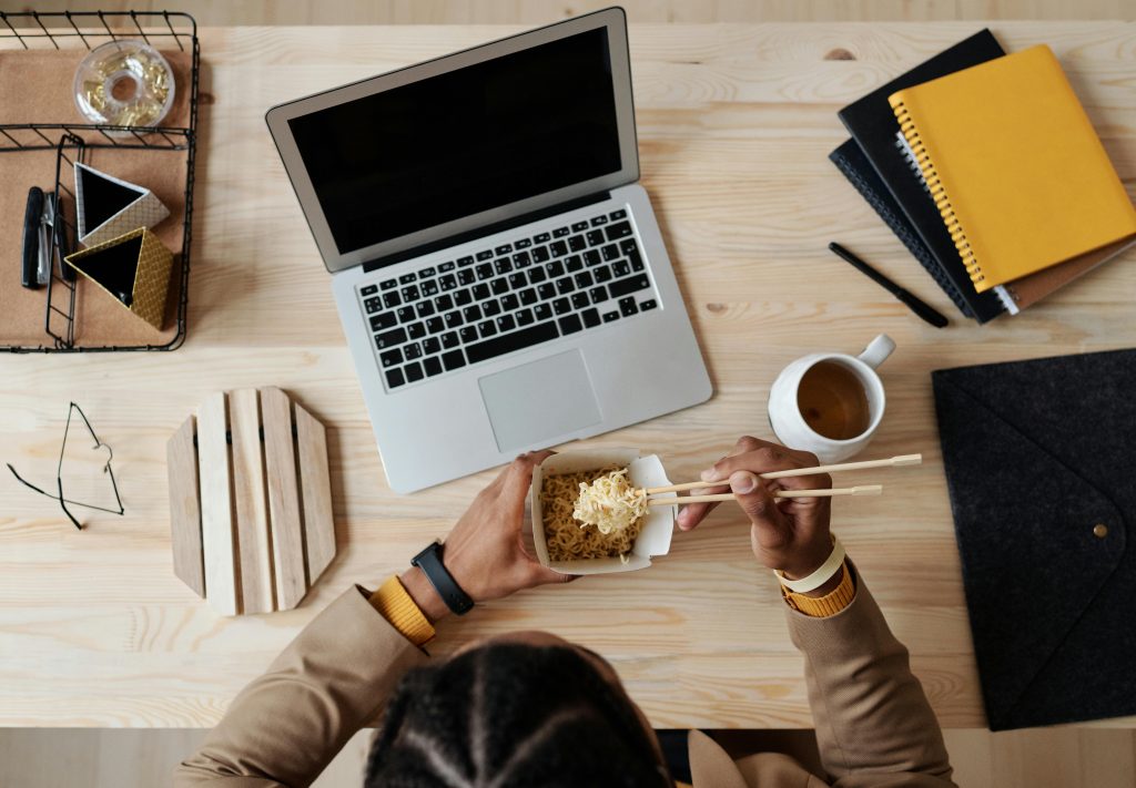 Persona comiendo fideos con palillos frente a una laptop en un escritorio de oficina.