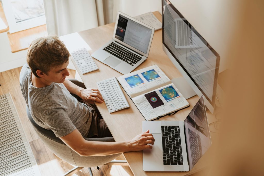 Persona trabajando en teletrabajo con dos portátiles, un monitor y un cuaderno abierto en un escritorio, mostrando una configuración de trabajo remoto.